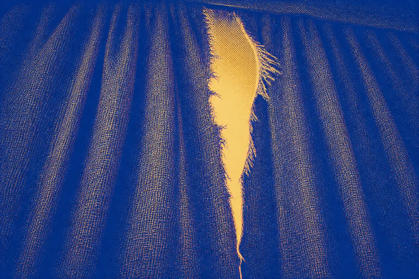 A single heavy woven temple curtain filling the frame, ripped open by one jagged tear that is widest at the top edge and tapers downward, frayed threads bristling along both torn edges, with warm light showing through the gap.